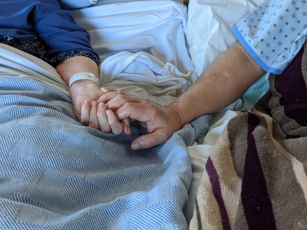 Always holding hands photo of two elderly people holding hands while lying in two hospitable beds pushed together.