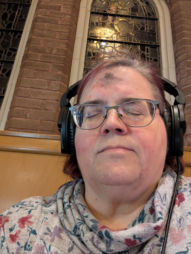 Photo of the author, a woman wearning glasses, and headphones, eyes are closed,and there is a smudge of ash on her forehead. Behind her is a brick wall and a darkend stained glass window.