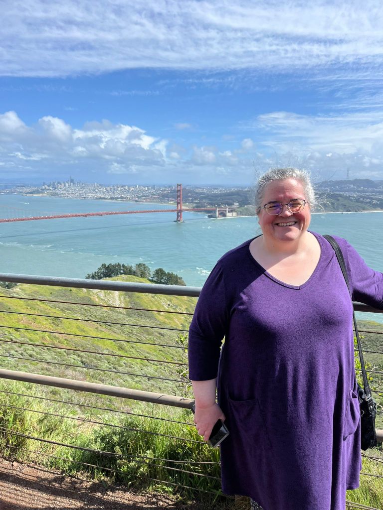 A photo of me, a woman with purple framed glasses, greying hair and a big grin, wearing a loose fitting purple tunic top, standing facing the camera with a view of the Golden Gate Bridge and the city of San Franscisco behind me.