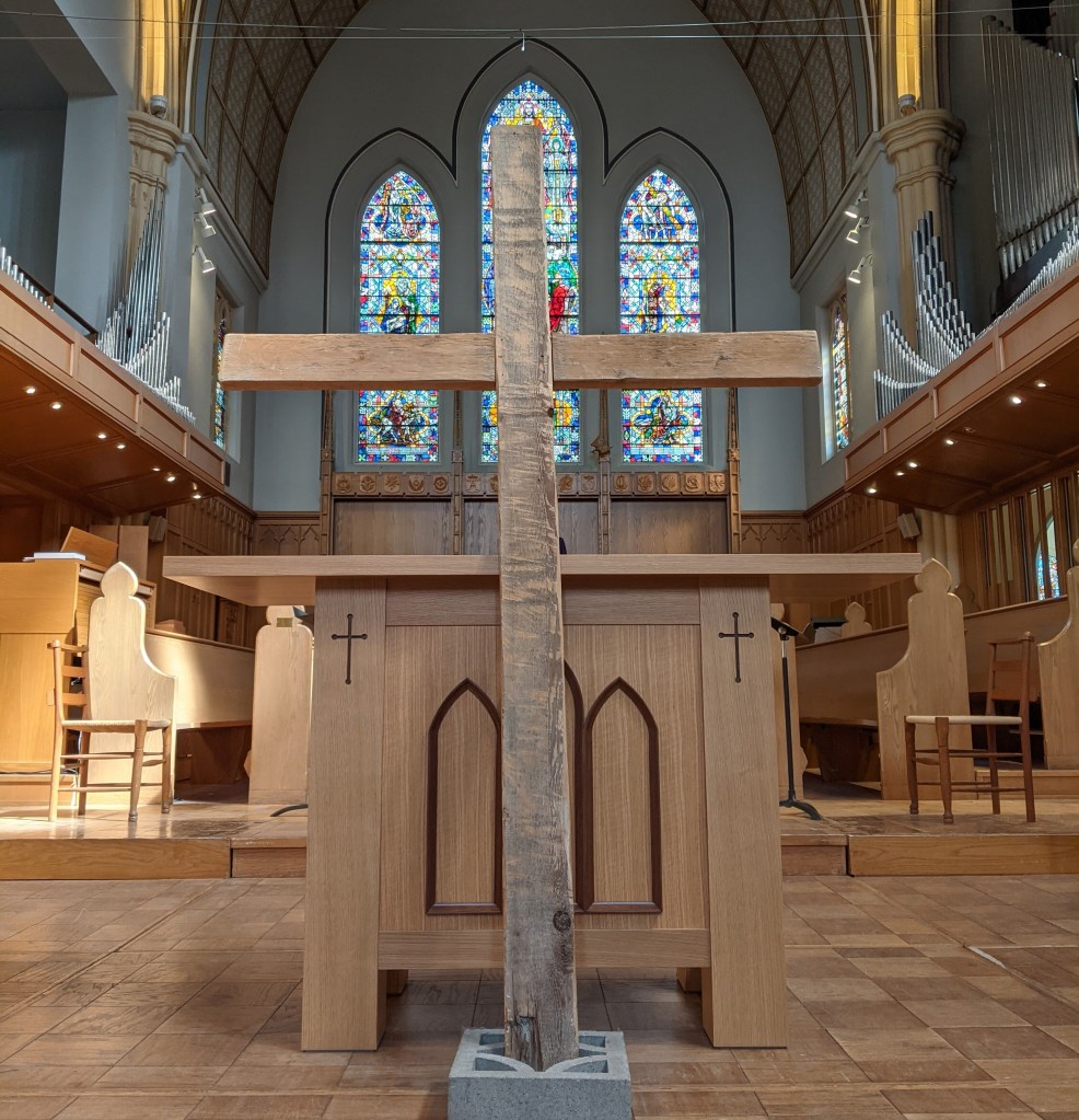 Photo of a very simple wooden cross in front of the alter with a trio of stained glass windows behind.