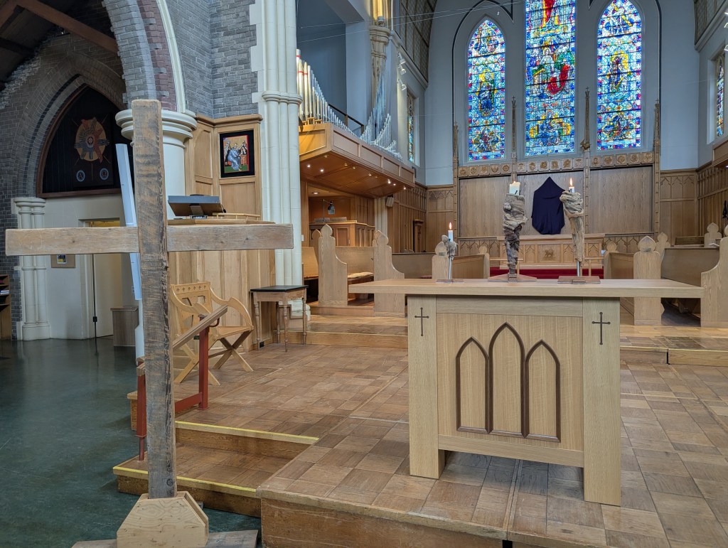 Photo of a simple wooden cross near the altar. On the altar sit three candles, made from driftwood on cross shaped bases. Behind the altar is a draped cross, an empty choir loft and three stained glass windows